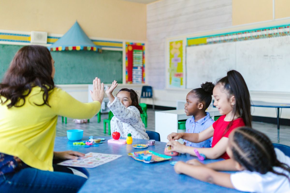 A teacher high-fives a smiling child in a colorful classroom with other children